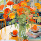Bright orange poppies in a glass vase on a table surrounded by books and a window backdrop, beautiful artwork from Garden Puzzle – Poppies Panorama: Orange Elegance – 1000 Piece Jigsaw Puzzle, 1000 piece jigsaw puzzles for adults.